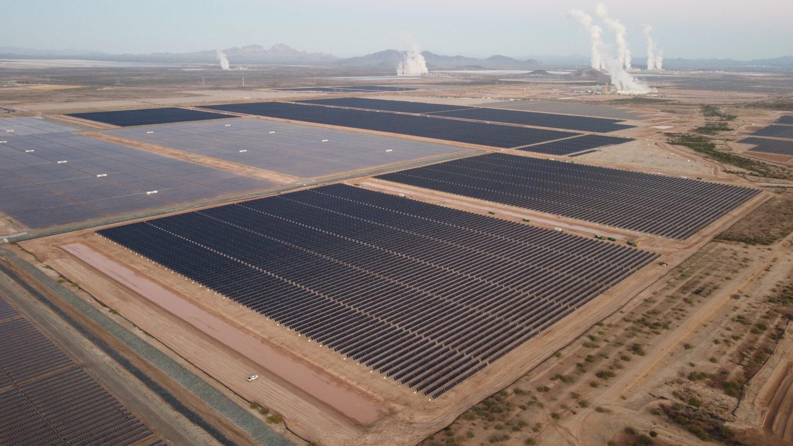 A desert landscape with solar grids in front of mountains with steam rising in the distance